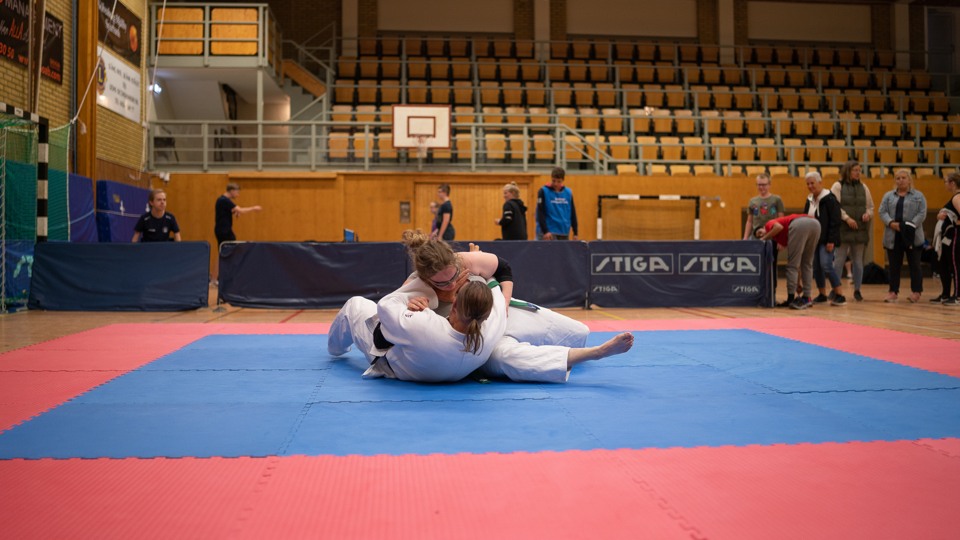 Två personer utövar judo på en röd golvmatta i en idrottshall.