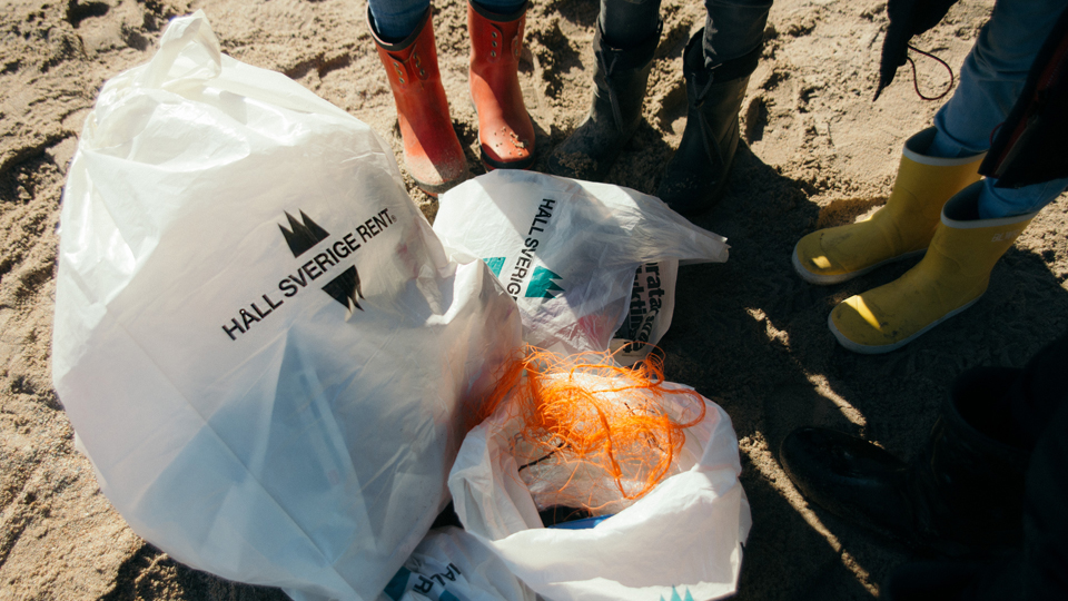 Fyllda skräppåsar med bland annat trasigt fisknät på en strand. På påsarna finns Sverige rents logotyp. I bakgrunden finns de personer som plockat skräpet.