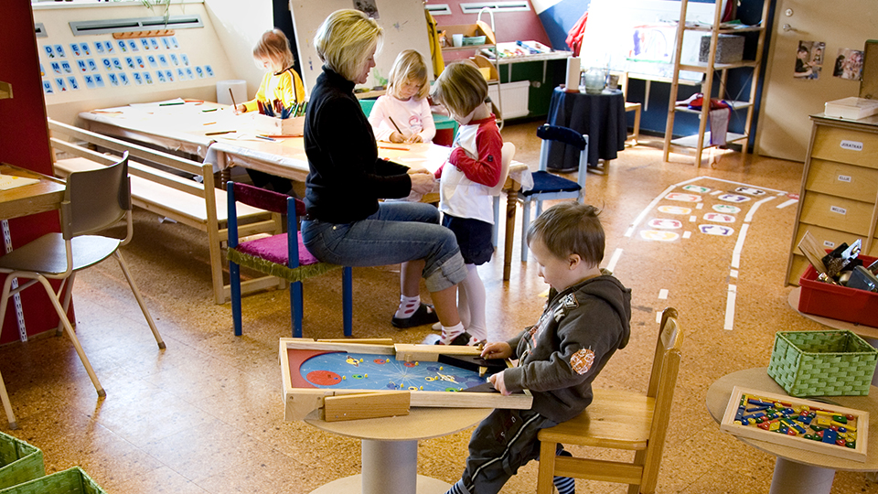En förskolepedagog sitter vid ett bord tillsammans med flera förskolebarn. De flesta barnen ritar och pysslar. 