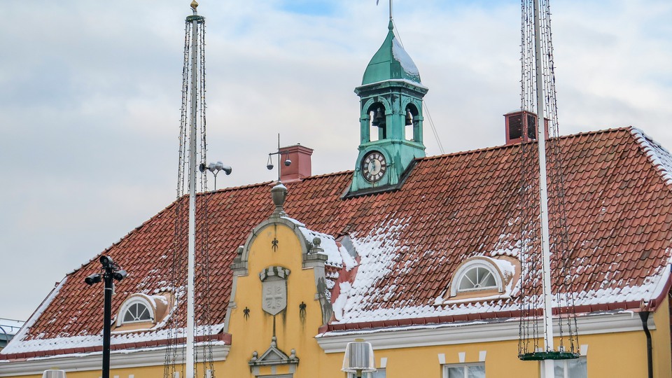 Snö ligger på taket på Sölvesborgs Rådhus, en vacker gul byggnad med ett klocktorn i koppar. Adventsljusstakar är tända i byggnadens fönster.