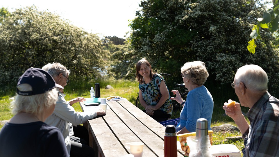 Bilden visar fem personer som sitter vid ett bord utomhus i grönska och fika