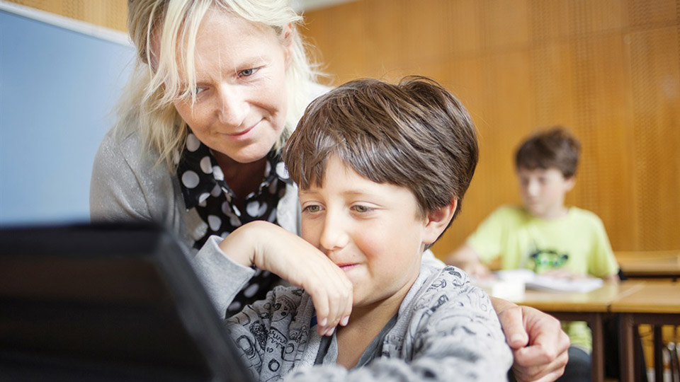 En lärare står bredvid en elev vid en bänk i ett klassrum. Båda tittar på en datorskärm. 