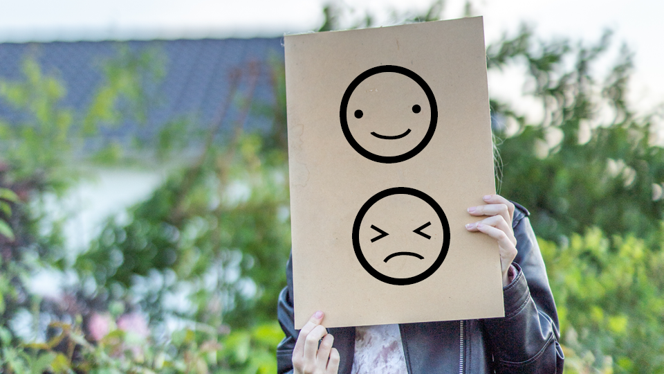 En flicka håller upp en brun pappkartong för ansiktet med illustrerade ansikten på, en glad gubbe och en sur gubbe.