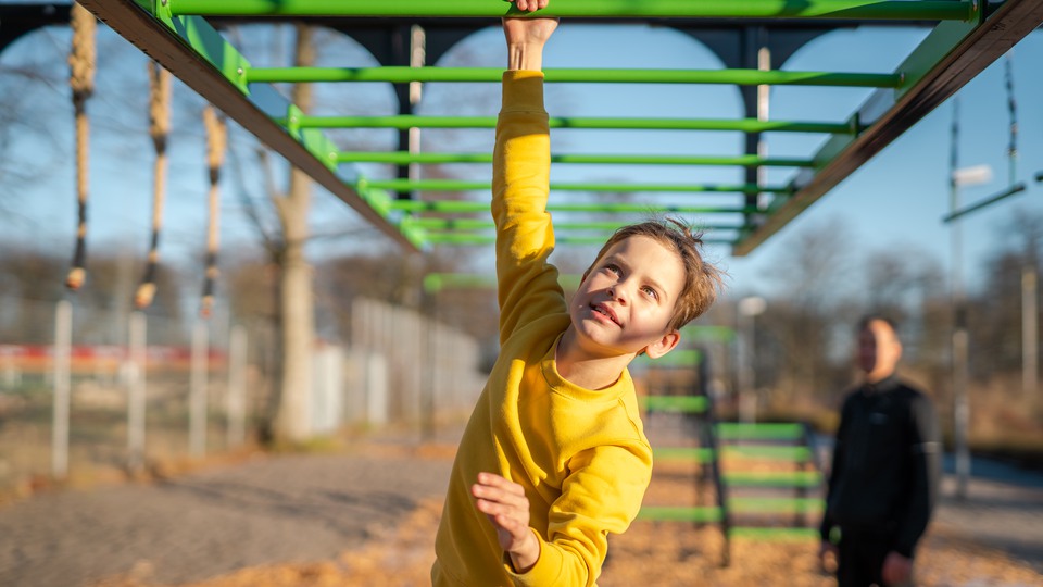 En ljust brunhårig pojke i saffransgul tröja svingar sig fram med händerna, hängades i gröna ribbor, i en OCR-bana en solig höstdag