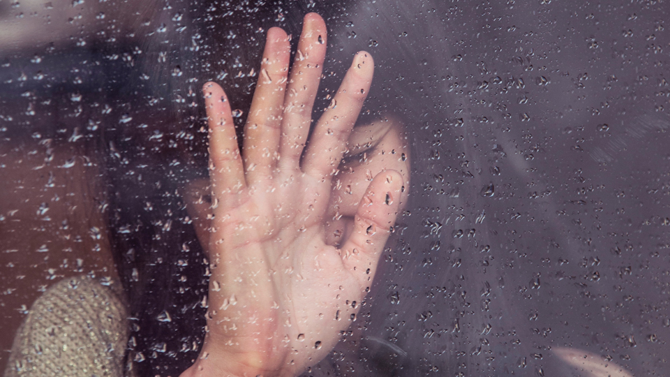 Bilden visar en anonym kvinnas hand mot ett fönster. Det regnar ute
