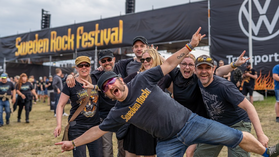 En grupp med människor som står framför huvudentré till Sweden Rock Festival. En person står på ett ben framför de andra. Alla är rockklädda och ser glada ut. 
