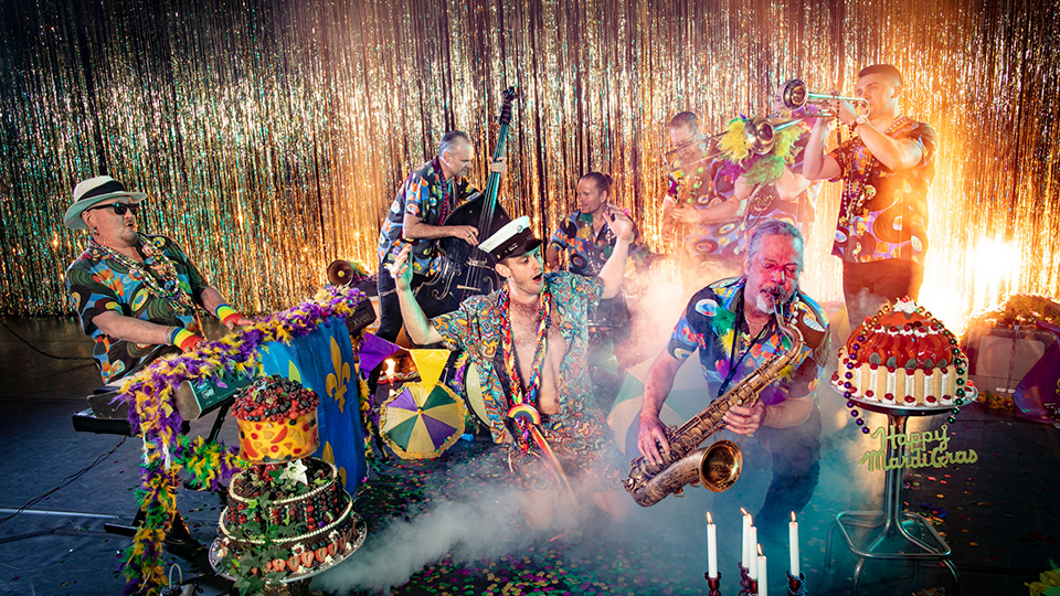 Bandmedlemmar i färgglada kläder och fjäderboas spelar framför ett guldigt draperi