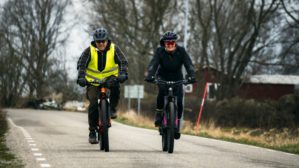 Ett äldre par cyklar på motionscykel på en landsväg och gör tummen upp.