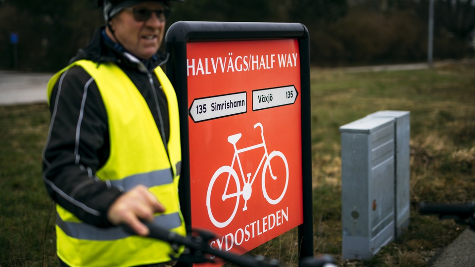 En man iklädd gul reflexväst och cykelhjälm står intill en röd skylt som det står "Halvvägsskylten, Sydostleden" på.