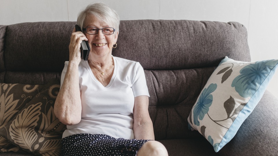 En äldre kvinna med kortklippt hår sitter i sin soffa klädd i vit t-shirt. Hon ser glad ut och pratar i telefon.