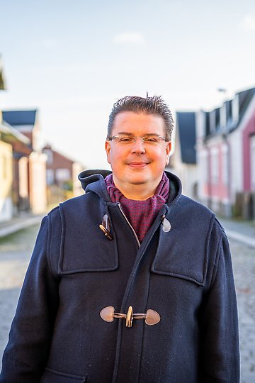 Marcus är klädd i mörkblå kappa och står ute på en kullerstensgat i Sölvesborg en solig vacker höstdag.
