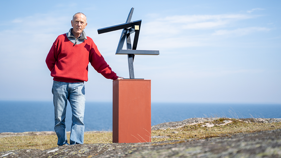 En äldre man, Johnny Martinsson klädd i röd tröja och blå jeans står på en kulle på Hanö med sin metallskulptur ur serien ”Drömda vinklar”.