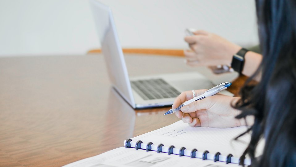 Person håller i penna över ett block medan man ser händer över en laptop i bakgrunden.