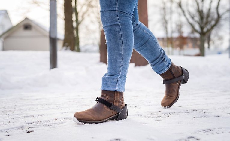 Bilden visar en person med halkskydd på sina skor i ett vinterlandskap.