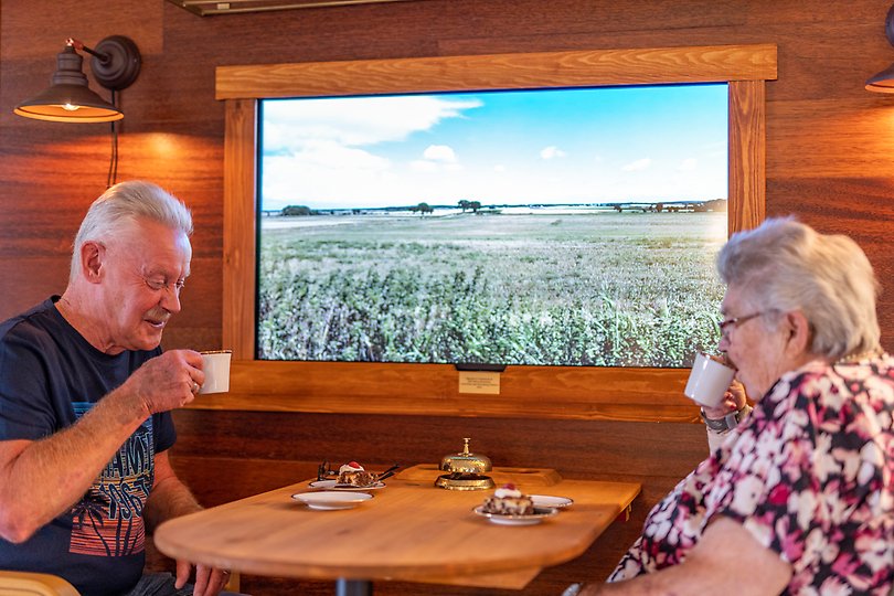 En äldre dam och herre sitter och dricker kaffe och tittar på en TV-skärm där de färdas över öppna jordbruksfält.