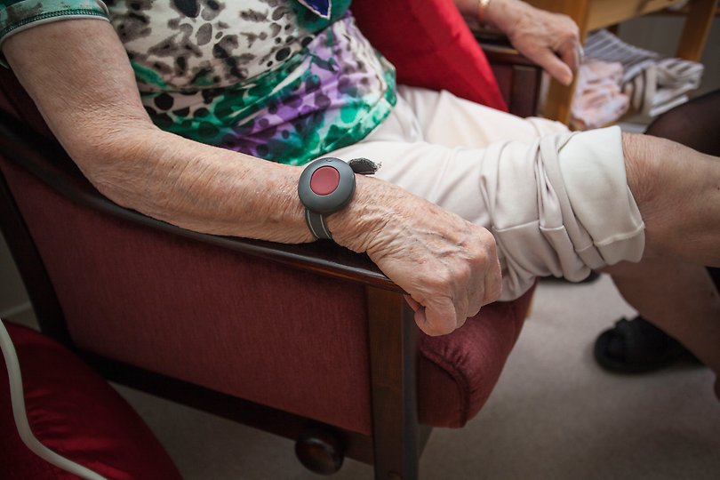 En äldre människa sitter i en fåtölj med sitt trygghetslarm på handleden. 