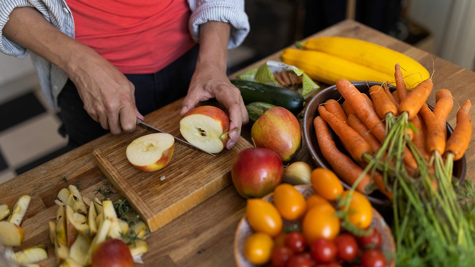 Skärbräda med frukt och grönsaker, händer som håller i en kniv och skär ett äpple.